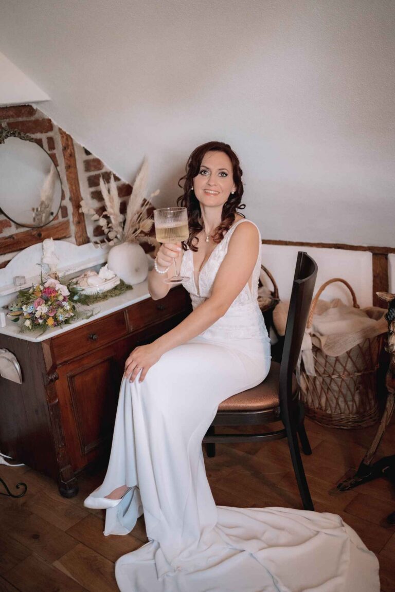 Braut sitzt lächelnd mit einem Glas Sekt in der Hand in einem gemütlichen Zimmer