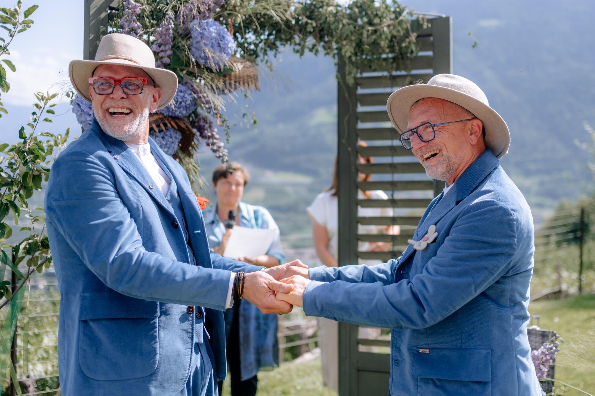 Thorsten Rausch und Ingmar Laimer halten sich lachend an beiden Händen vor dem Traubogen ihrer freien Trauung in Südtirol